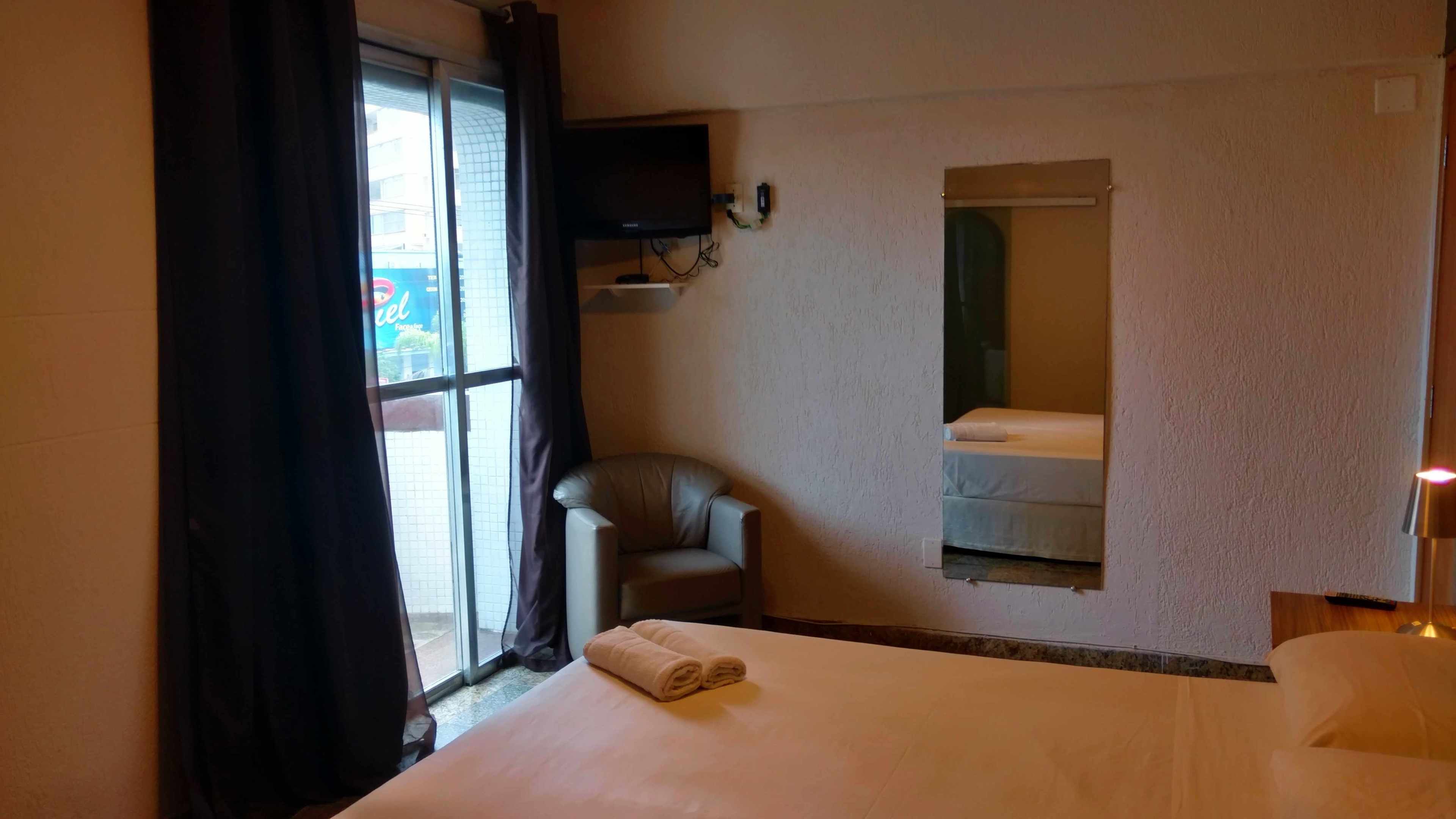 A hotel room with a bed, a chair, a television mounted on the wall, and a window with curtains.