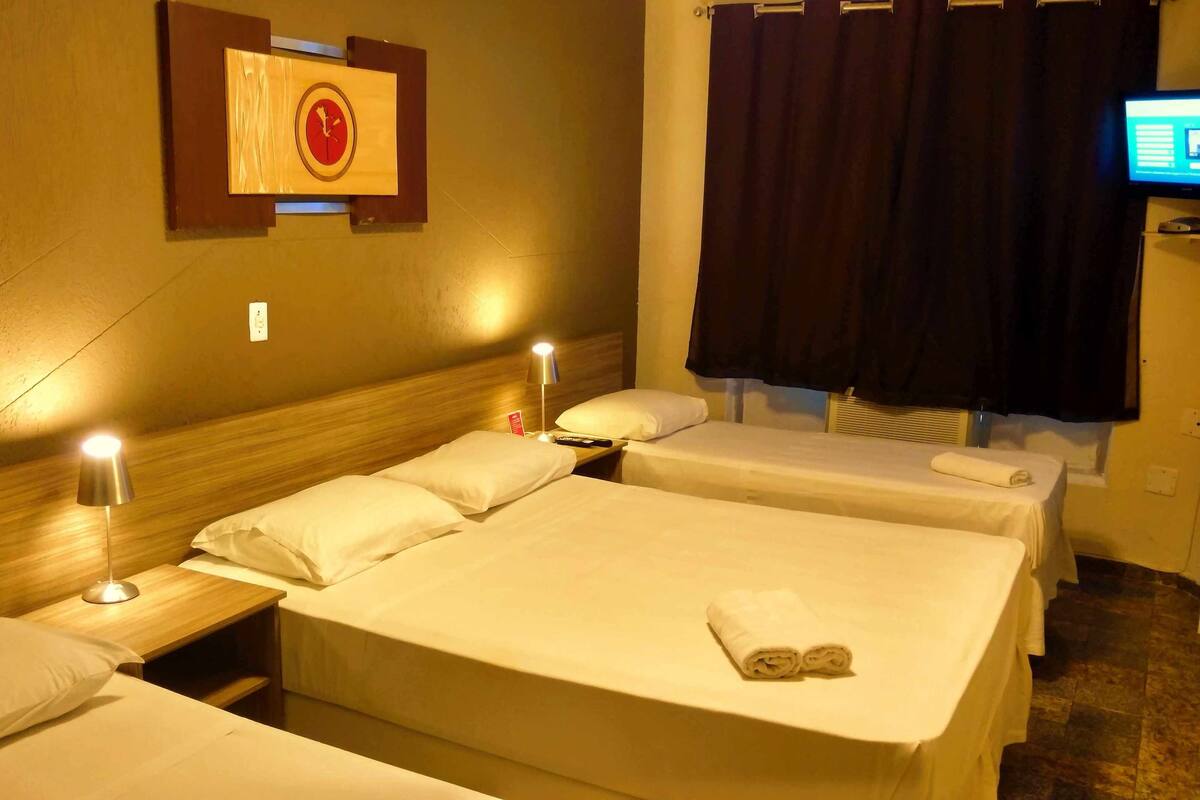 A hotel room with two beds, a nightstand with a lamp, a wall clock, a television, and a window with curtains.