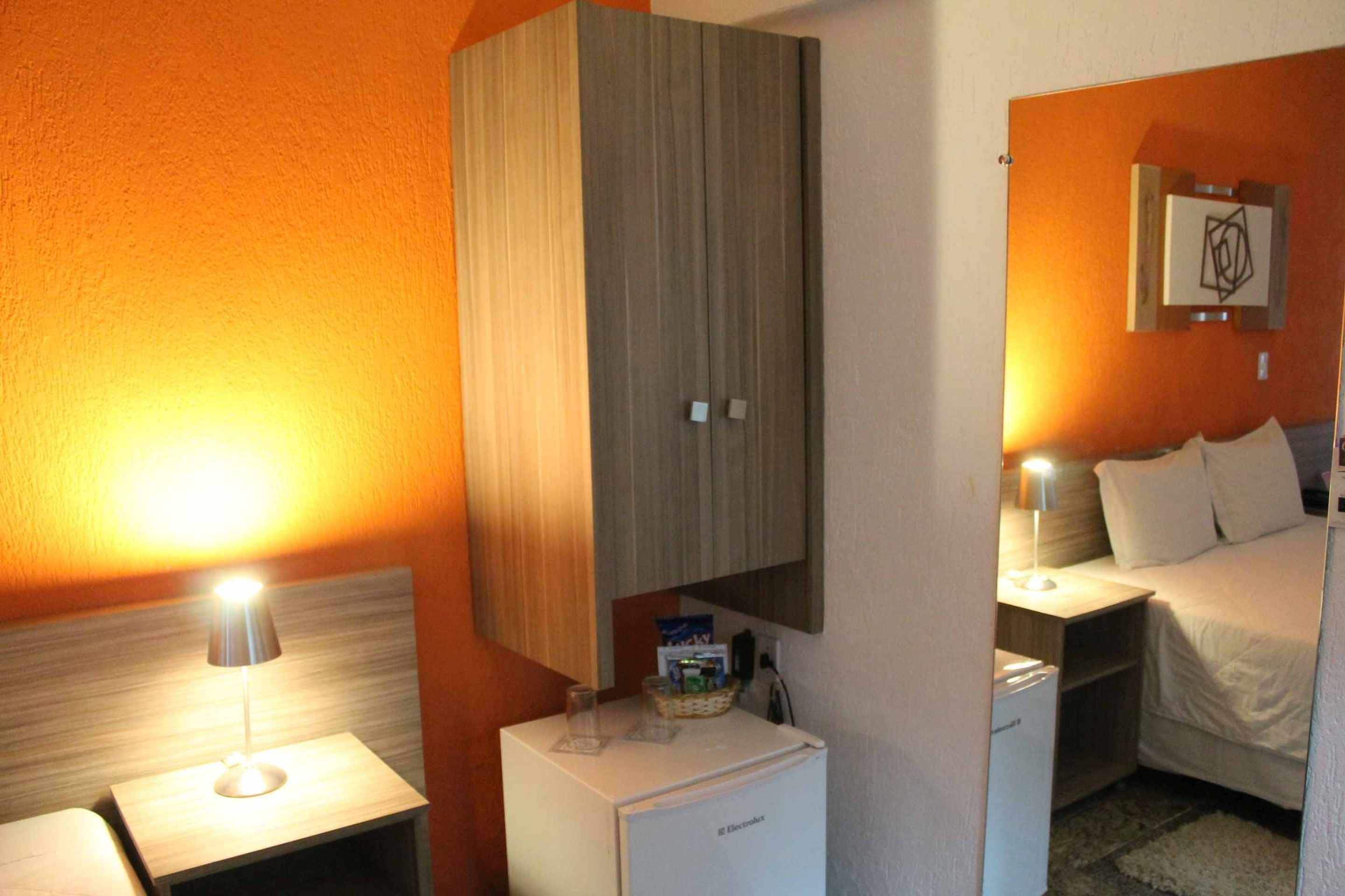 Una habitación de hotel con una cama, una mesita de noche con lámpara, un refrigerador pequeño y un armario de madera.