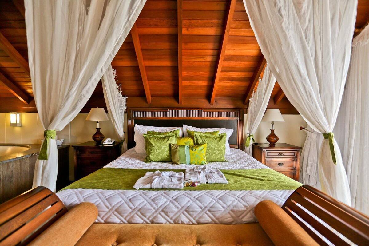 A bed with a wooden headboard, white and green bedding, and a canopy. There are two bedside tables with lamps and a nightstand with a phone.