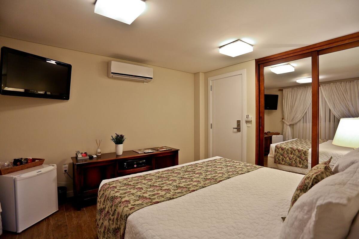 A hotel room with a bed, a desk with a computer, a small refrigerator, an air conditioner, and a television on the wall.