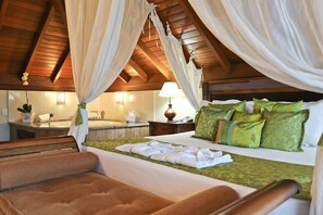 A bedroom with a canopy bed, a sitting area with a cushioned bench, a bedside table with a lamp, and a bathroom with a sink and mirror.