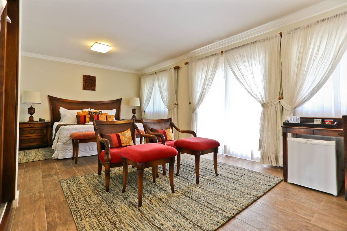A hotel room with a bed, two armchairs, a small table, a refrigerator, and a window with curtains.