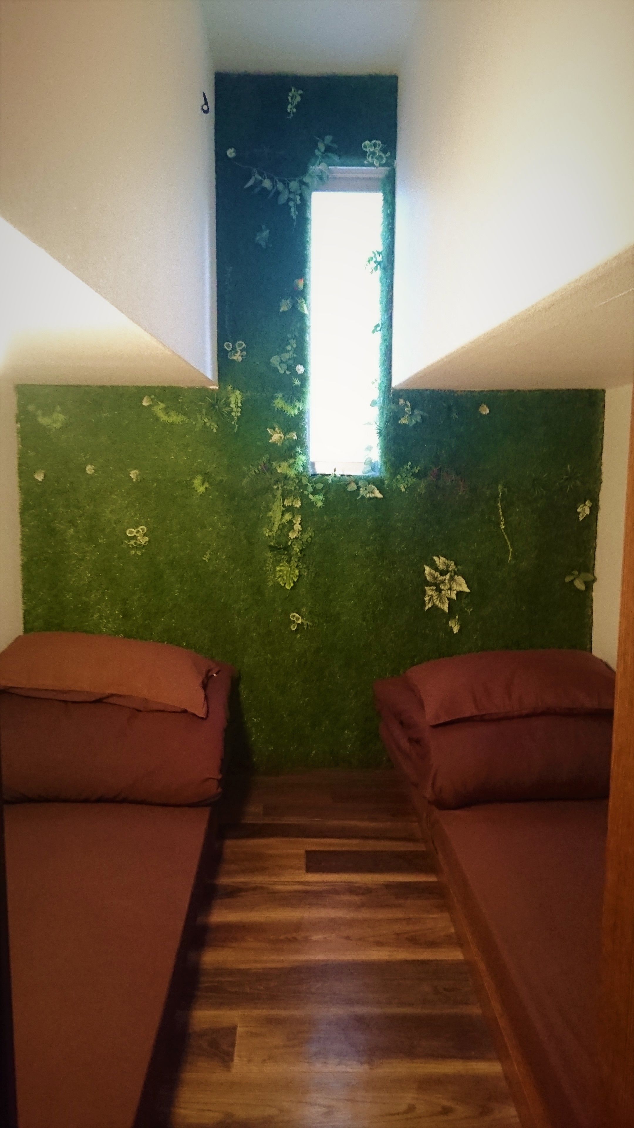 A room with two beds, a green wall with floral patterns, a small window, and a wooden floor.
