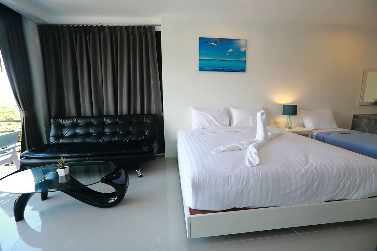 A modern hotel room with a large bed, a glass coffee table, a black leather sofa, and a view of the sea through the window.