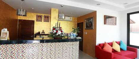 Un área de recepción con un mostrador revestido de mosaicos, un sofá rojo con cojines de colores y un amplio ventanal que proporciona luz natural.
