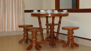 Una mesa redonda de madera con cuatro taburetes a juego, una mesita auxiliar con una tetera y tazas, y una ventana con marco de madera.