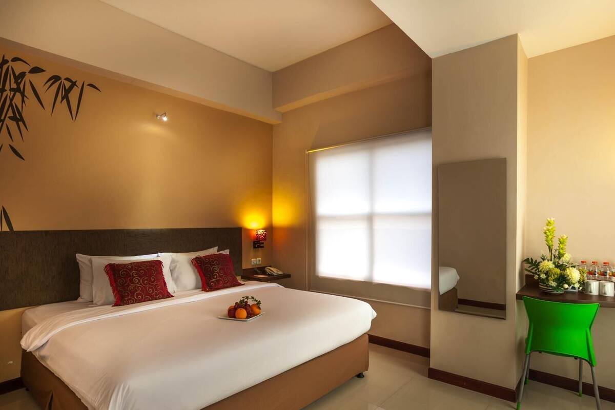 A hotel room with a large bed, a bedside table with a lamp, a green chair, and a window with blinds.