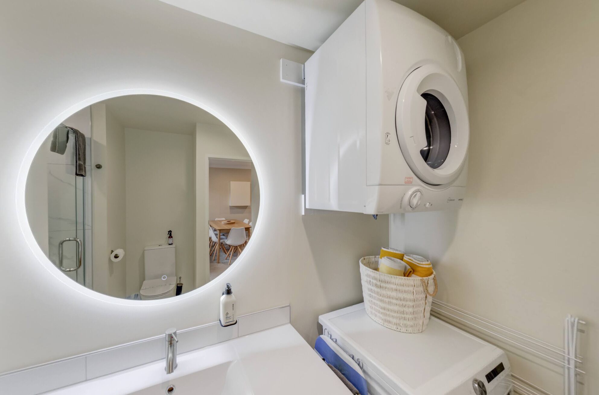 A modern bathroom with a round mirror, a built-in washing machine, and a small basket with towels.