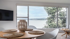 A dining area with a large window offering a view of a body of water and trees. - Harbour View Motel and Water Front Apartments (Tauranga)