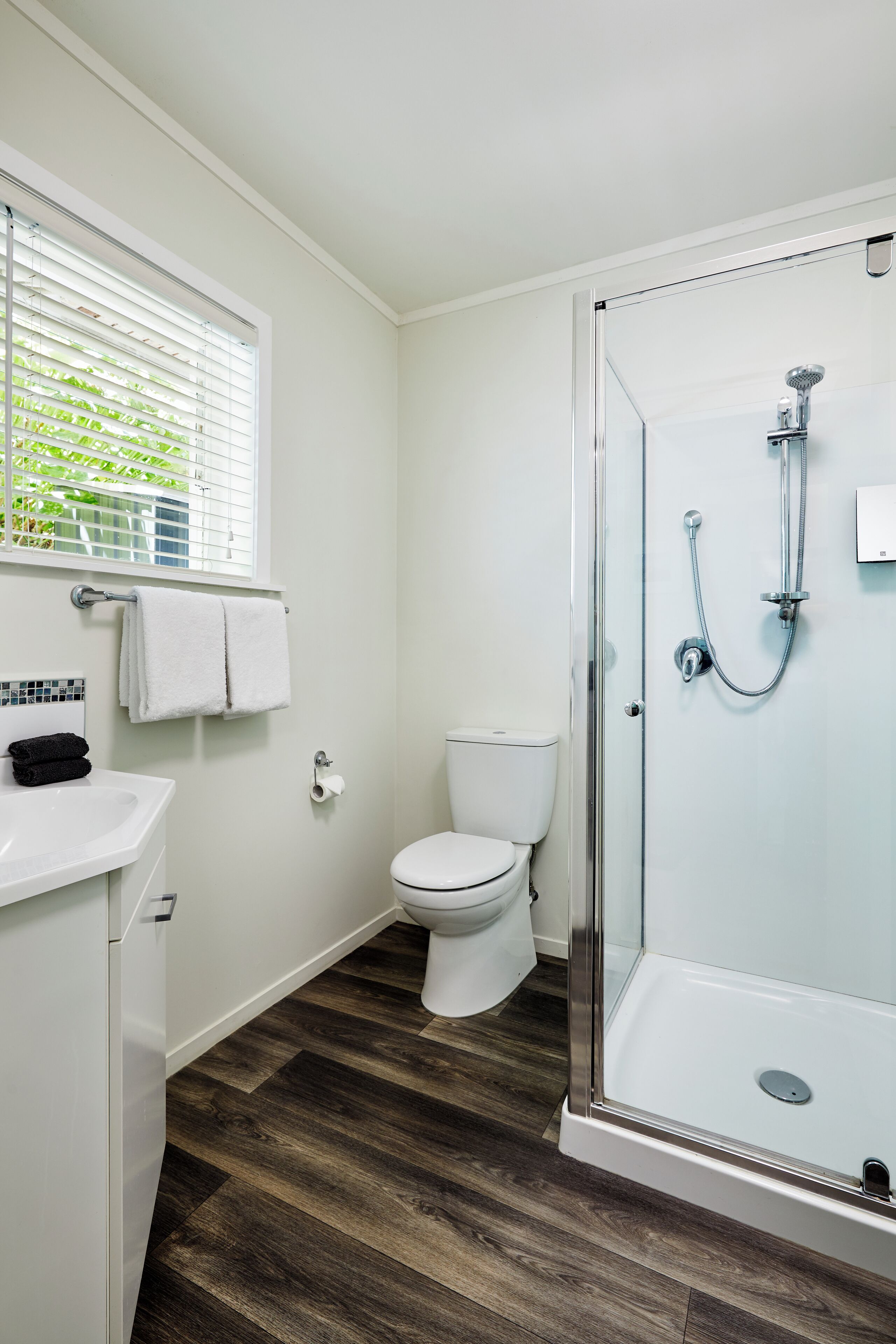Ein Badezimmer mit Duschkabine, weißer Toilette, Waschbecken mit Spiegel und Fenster mit Rollo.