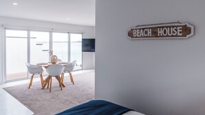 A modern bedroom with a large window, a dining area with a table and chairs, and a sign on the wall that reads "BEACH HOUSE". - Harbour View Motel and Water Front Apartments (Tauranga)