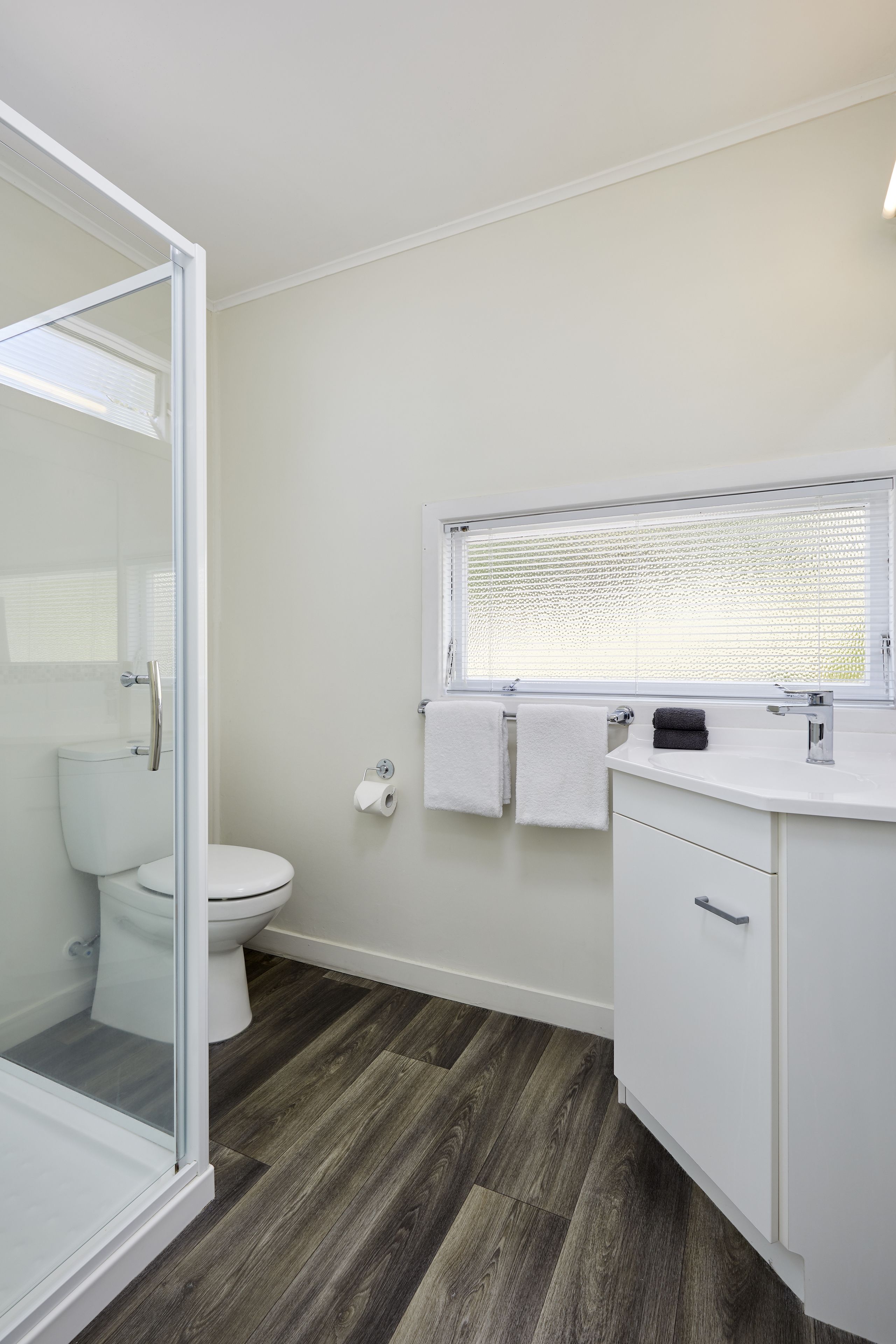 Een badkamer met een douchecabine van glas, een wit toilet, een wastafel met een raam erboven en een donker houten vloer.