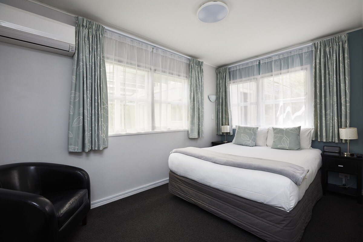 Habitación de hotel con cama, una silla, mesita de noche y ventana con cortinas.
