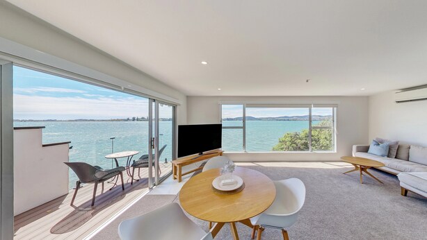 A modern living room with a large sliding glass door leading to a balcony overlooking a body of water. - Harbour View Motel and Water Front Apartments (Tauranga)