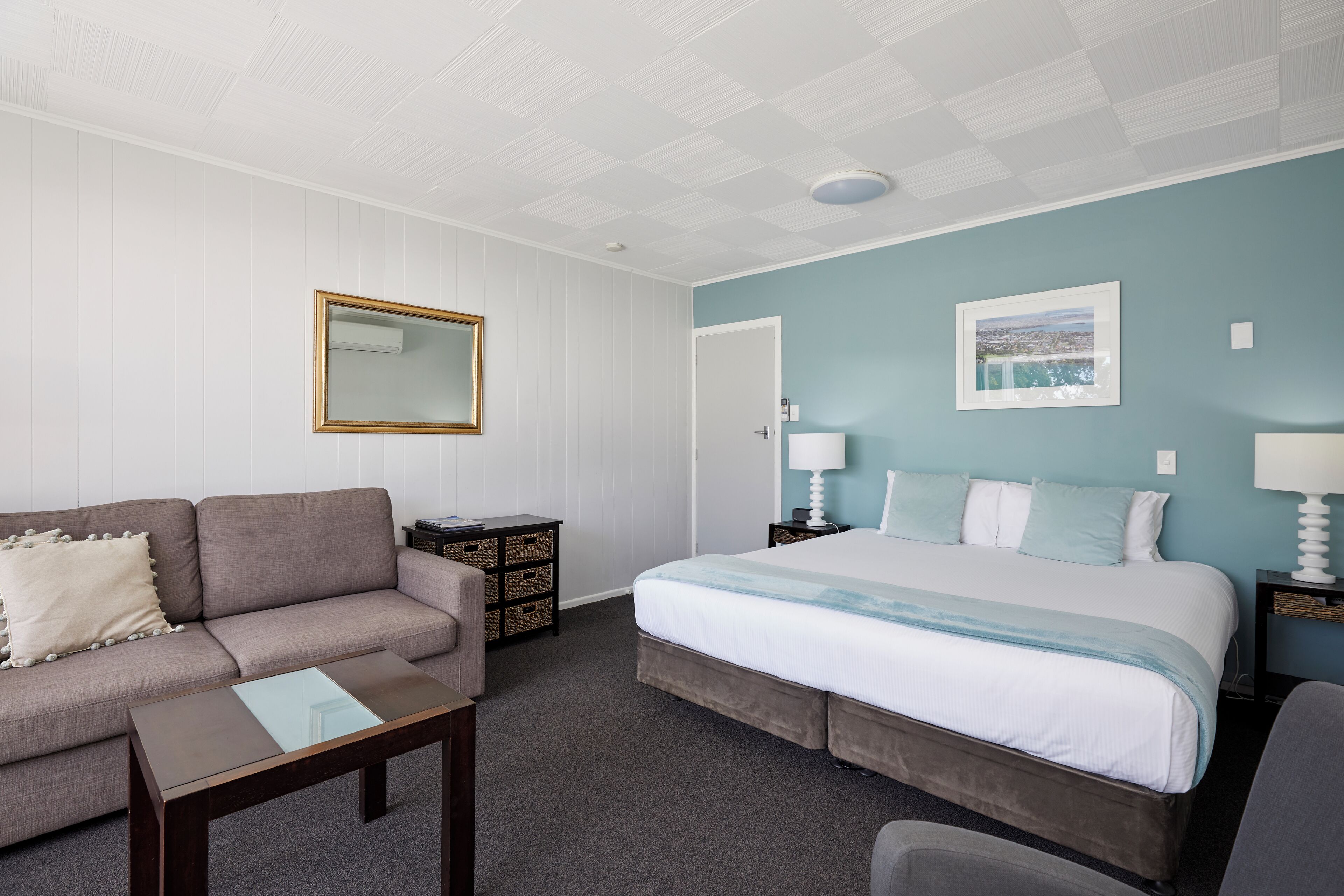 A hotel room with a bed, a sofa, a small table, a chair, a nightstand, and a wall-mounted mirror.