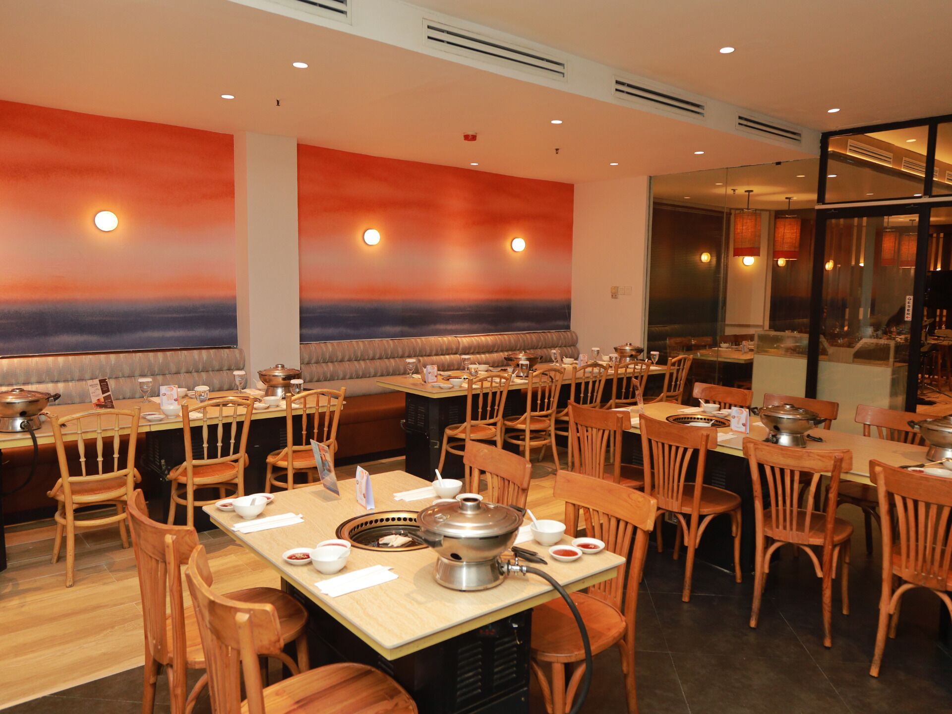 Un restaurante con mobiliario de madera, una disposición central para olla caliente y un mural de atardecer en la pared.