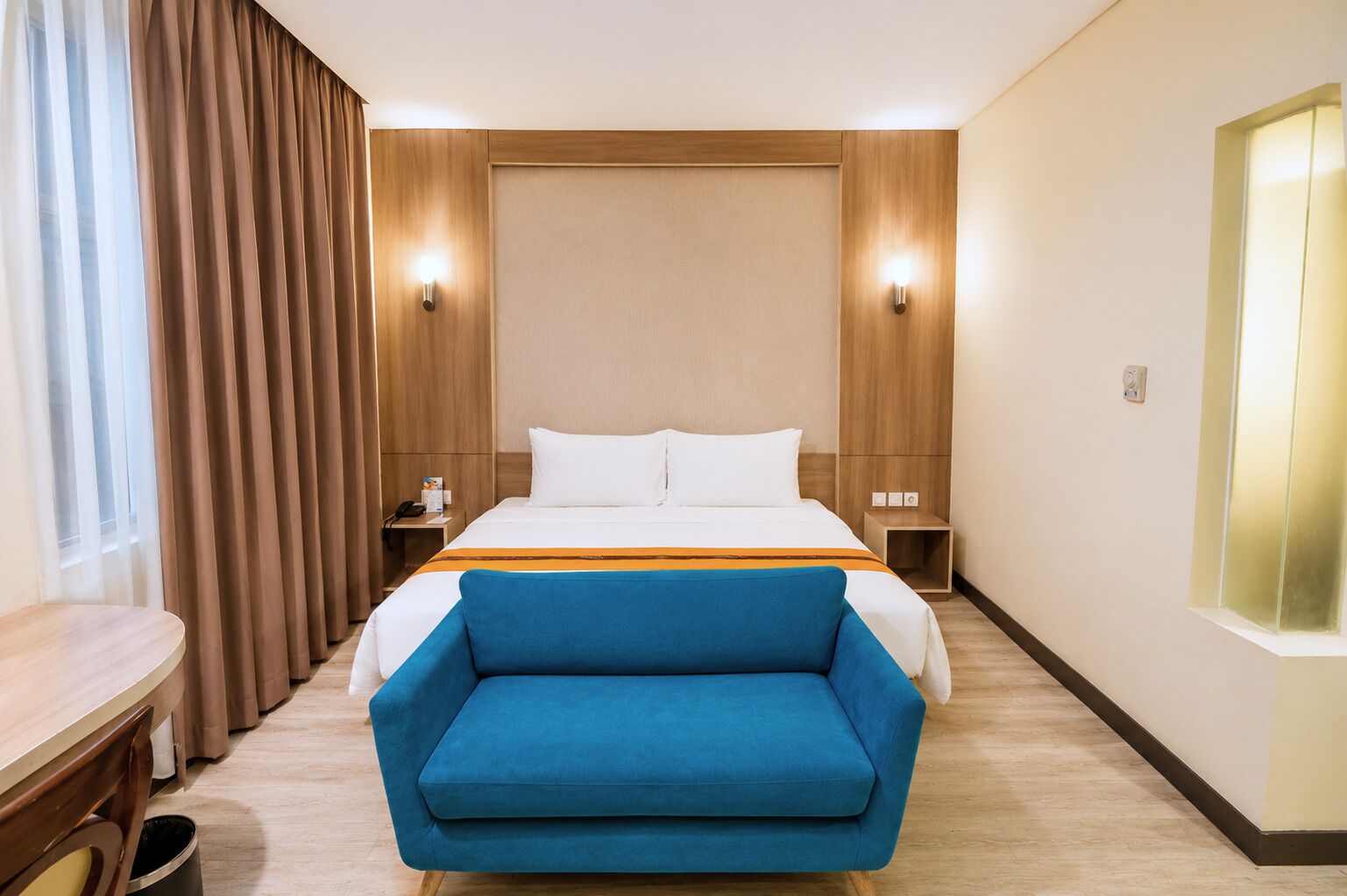 Habitación de hotel con cama, un sillón azul, una mesita de noche de madera, una mesita auxiliar y un aplique de pared.