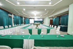 Un salón de conferencias con una larga mesa dispuesta para una reunión, rodeada de sillas blancas, y una pantalla de proyector al frente.