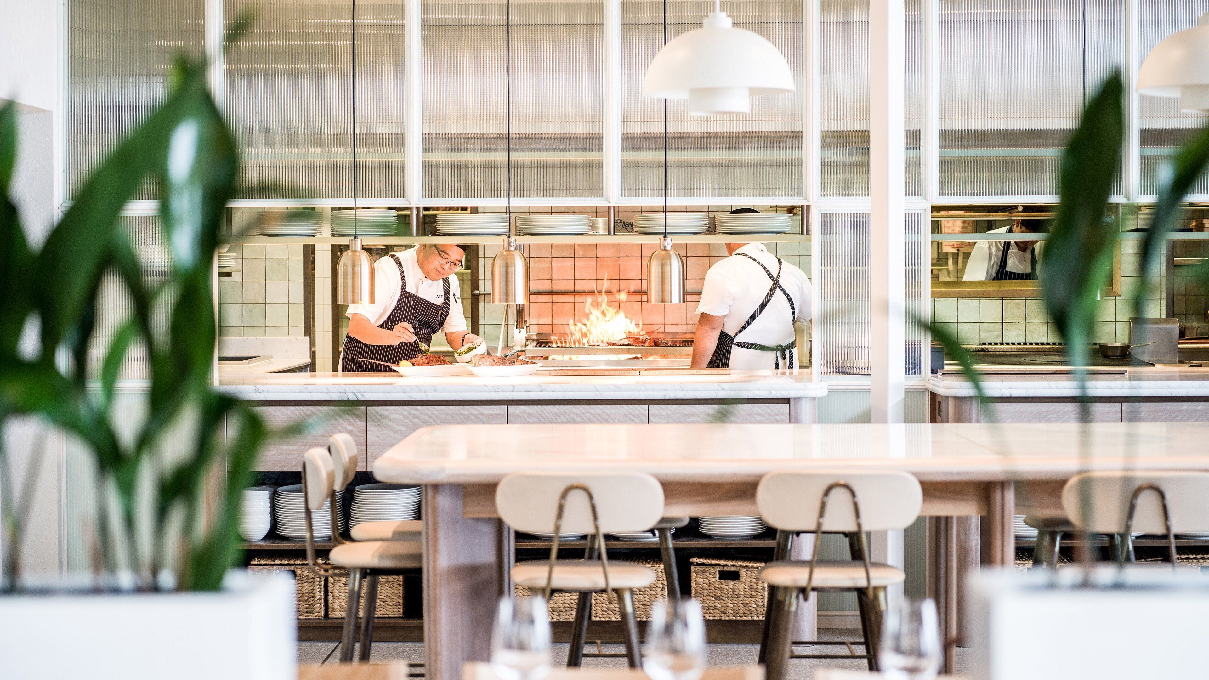 Un restaurante moderno con un área de cocina limpia y organizada, visible a través de un tabique de vidrio. Hay varias mesas y sillas dispuestas para los comensales.