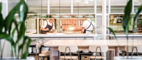 Un restaurante moderno con un área de cocina limpia y organizada, visible a través de un tabique de vidrio. Hay varias mesas y sillas dispuestas para los comensales.