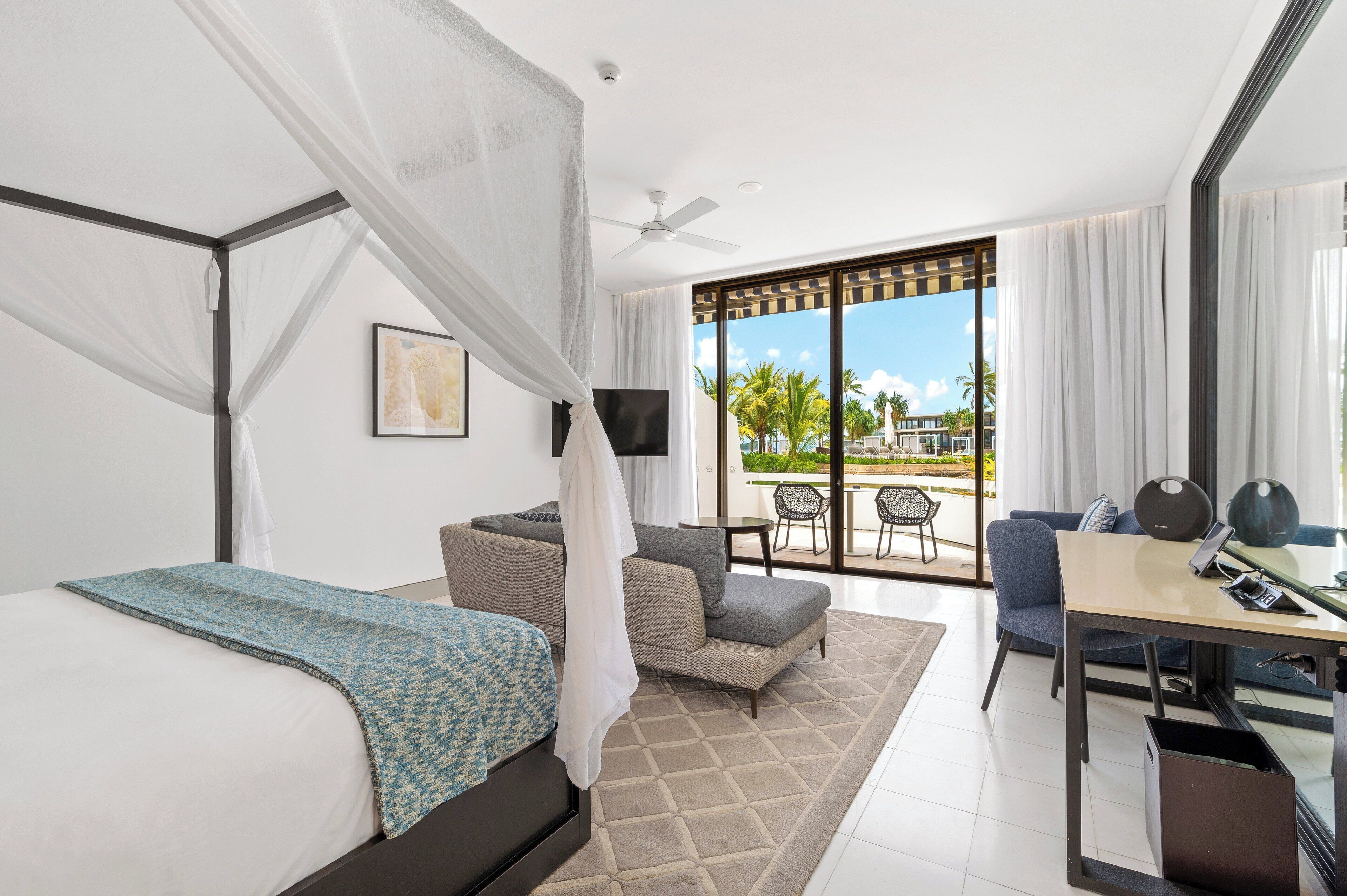 A modern hotel room with a bed, a sofa, a desk, and a balcony with a view of palm trees.