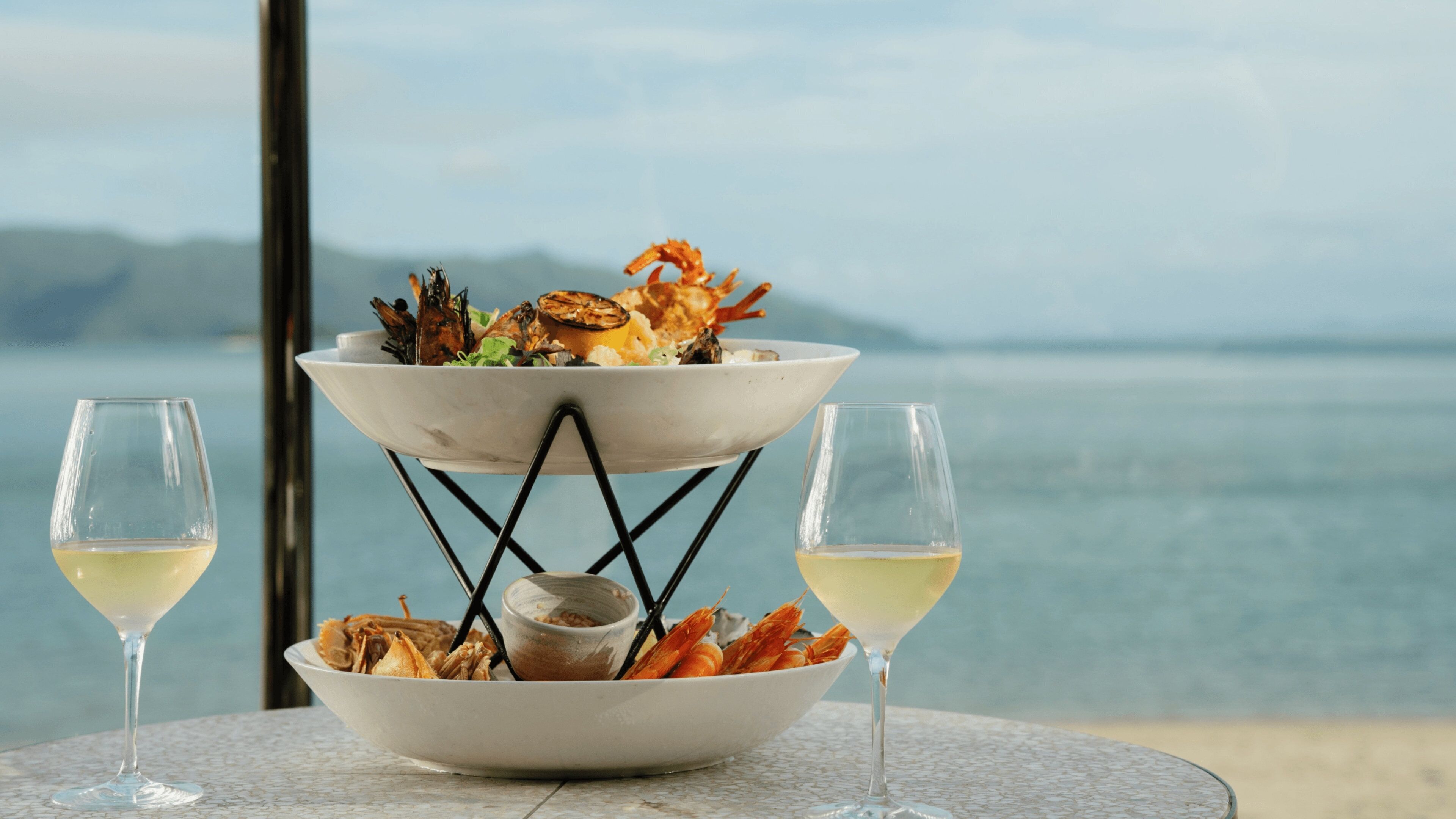 A seafood dish on a white stand with two glasses of white wine on a table by a window overlooking the sea.