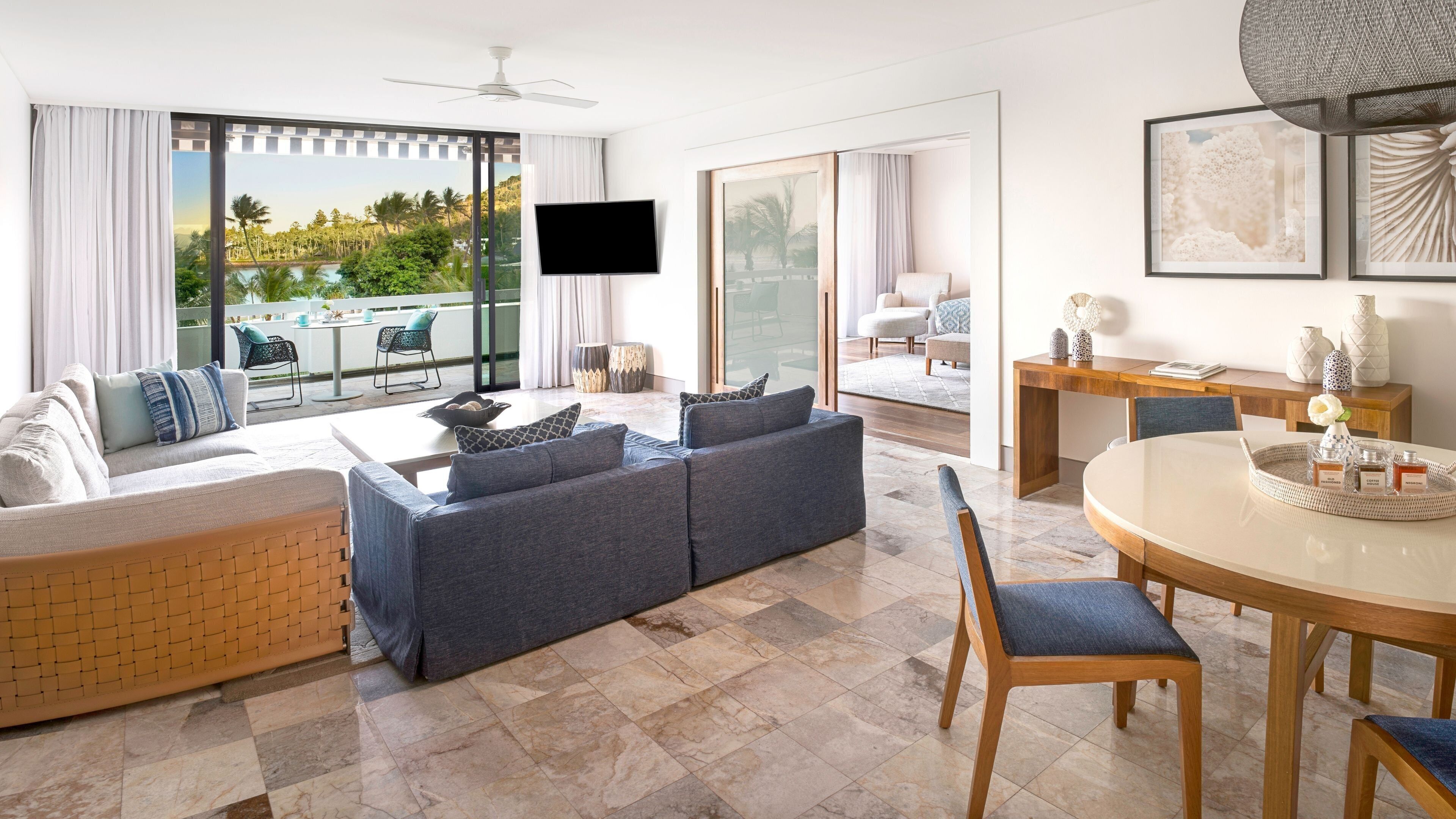 A modern living room with a large grey sofa, a dining table, and a view of a pool and greenery.