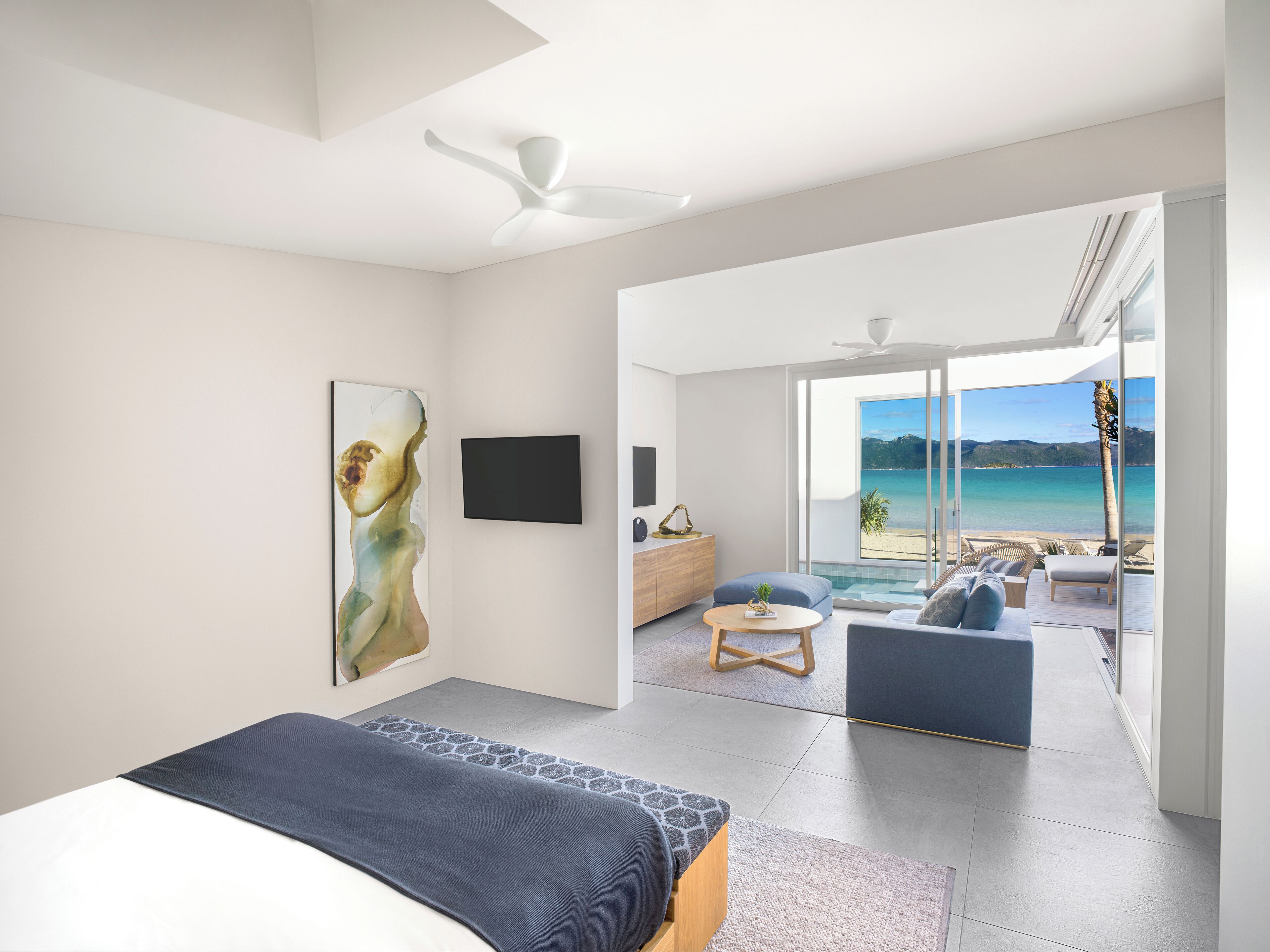 A modern bedroom with a large bed, a painting, a TV, and a balcony with a view of the sea.