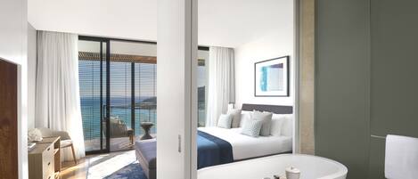 Une chambre d’hôtel moderne dotée d’un grand lit, d’une baignoire et offrant une vue sur la mer grâce à de grandes fenêtres du sol au plafond.