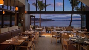 Ein Restaurant mit Tischen und Stühlen, das auf ein Gewässer mit Palmen und Bergen im Hintergrund blickt.