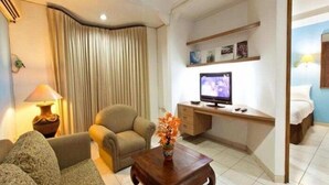 A hotel room with a sofa, a small table with a vase, a TV on a wooden stand, and a bed with a nightstand. - Maven Buncit (Jakarta)