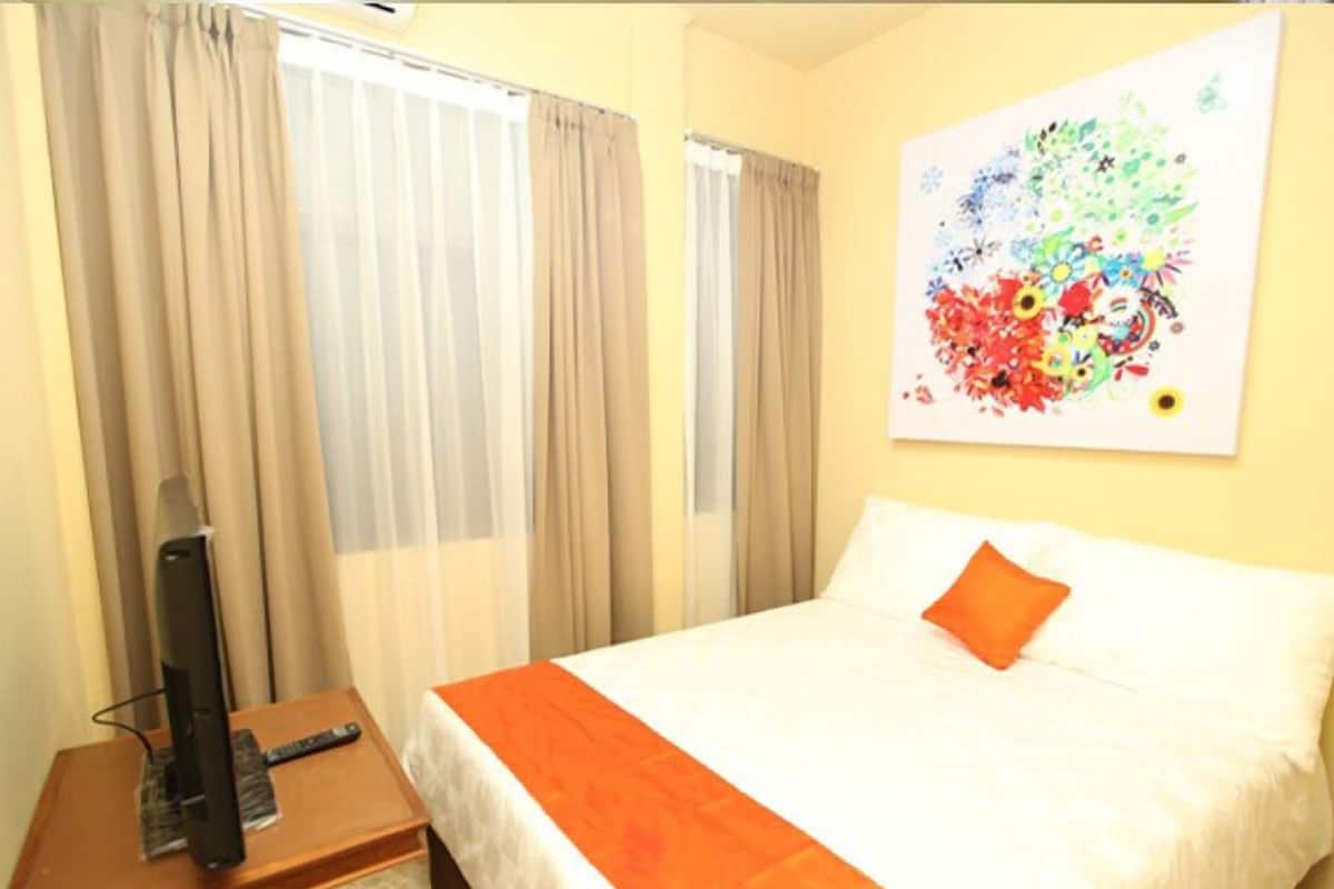 Ein Hotelzimmer mit einem Bett, einer orangefarbenen Tagesdecke, einem Fernseher auf einem Holztisch und einem farbenfrohen, abstrakten Gemälde an der Wand.