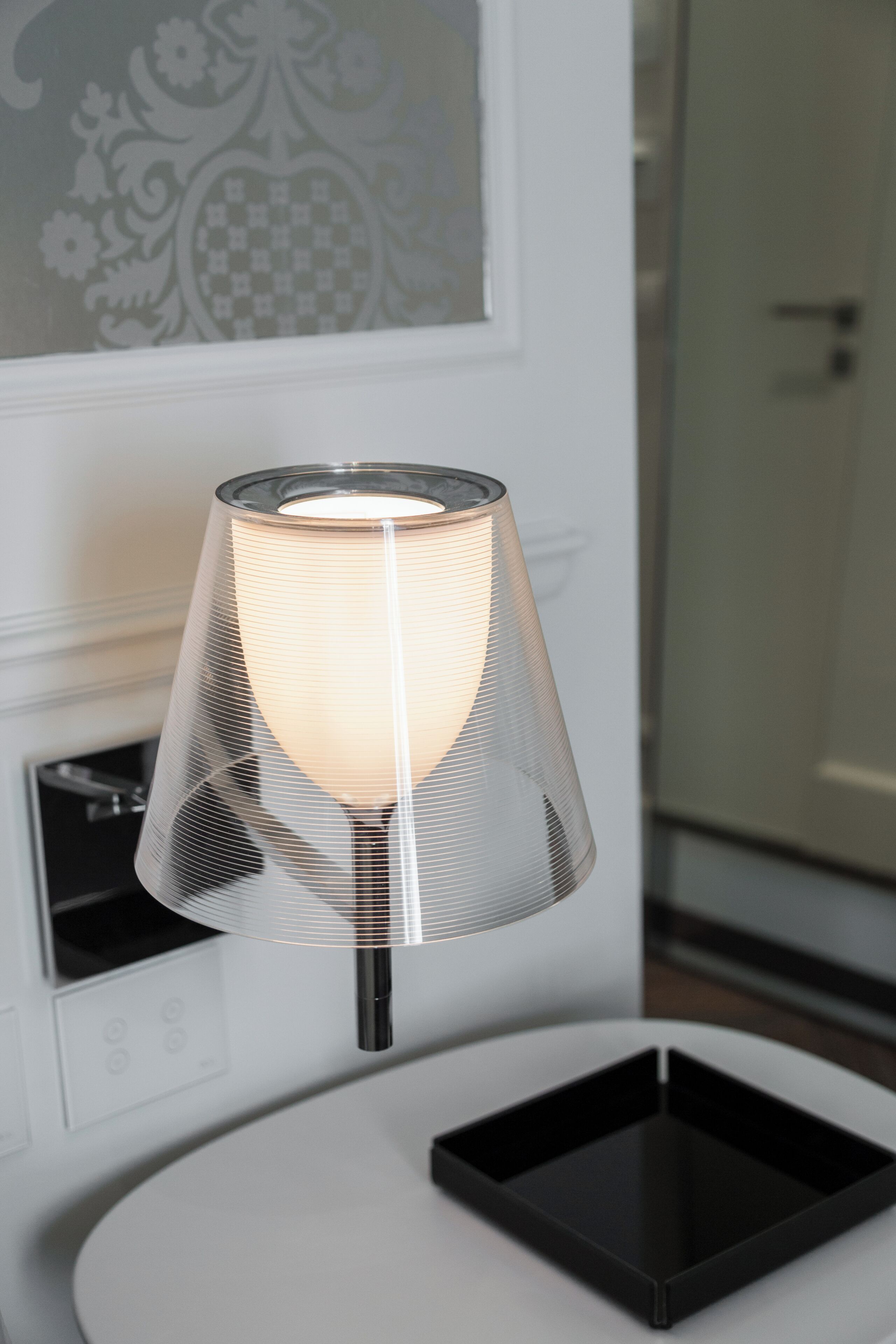 A modern lamp with a metallic shade on a white table in a room with a patterned wall and a door in the background.