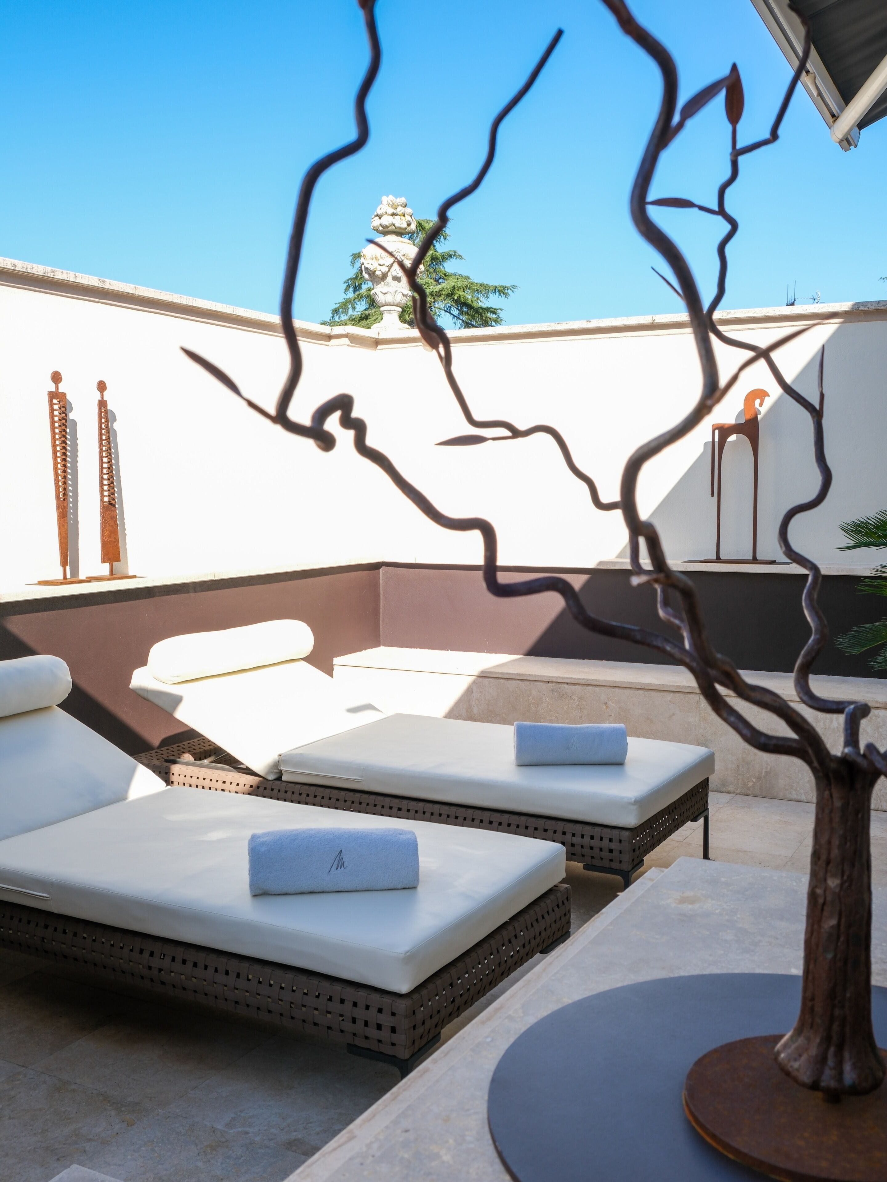 Un área de lounge en la azotea con sillones de sol, una escultura decorativa de árbol y una pared blanca con una escultura de metal ornamental.