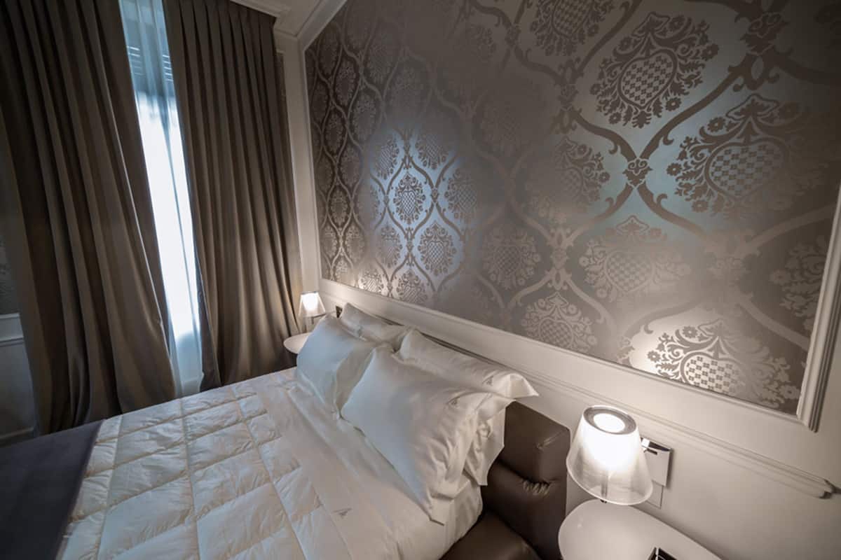 A hotel room with a large bed, a nightstand, a lamp, and a patterned wallpaper.