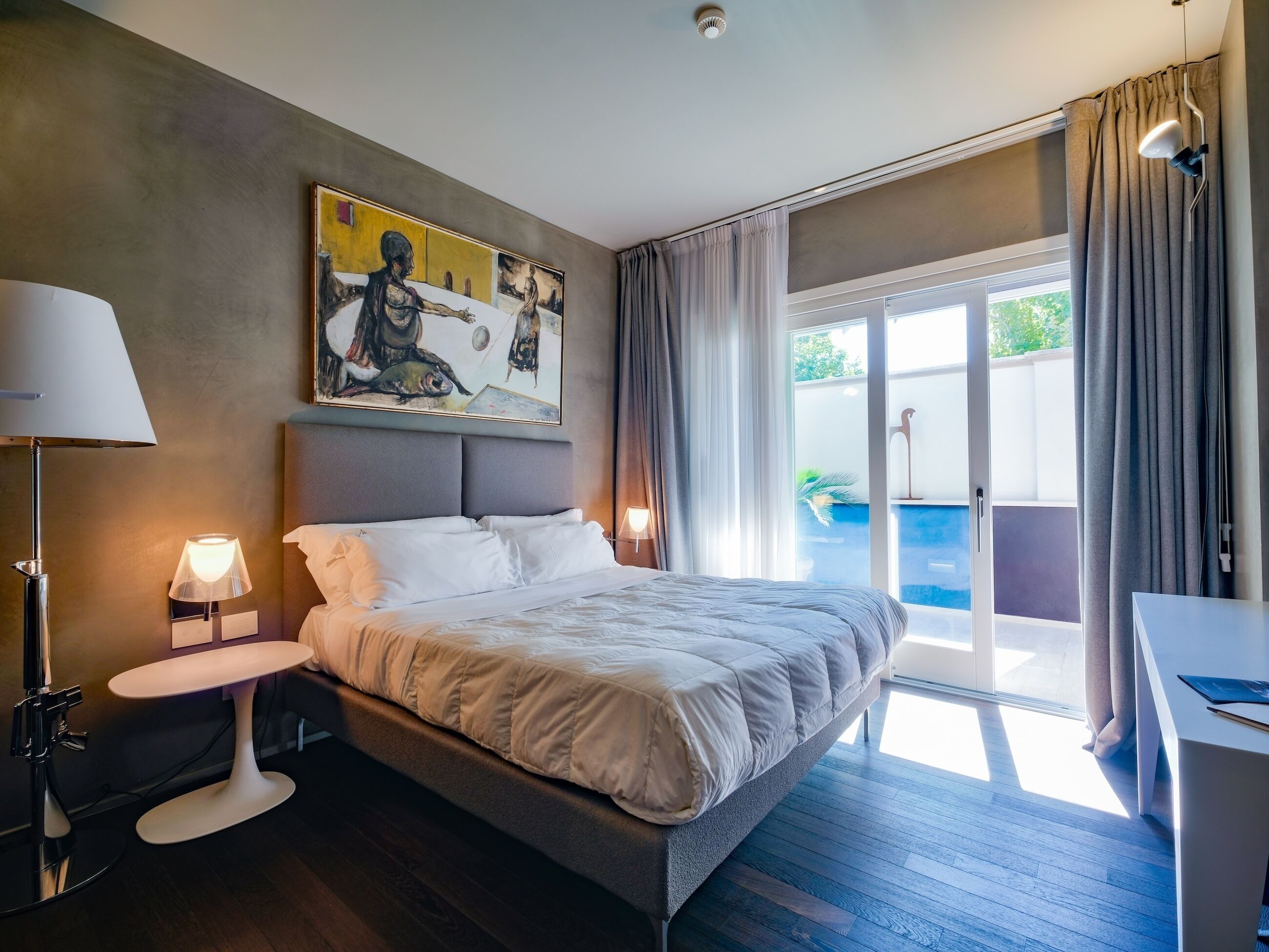 A modern hotel room with a large bed, a bedside table, a desk, and a painting on the wall.
