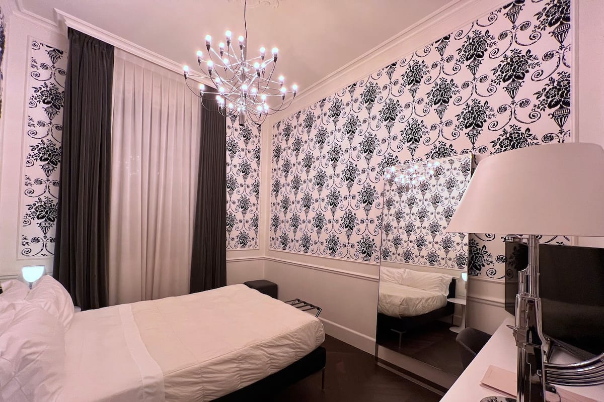 A hotel room with a bed, a mirror, a desk with a lamp, and a chandelier.