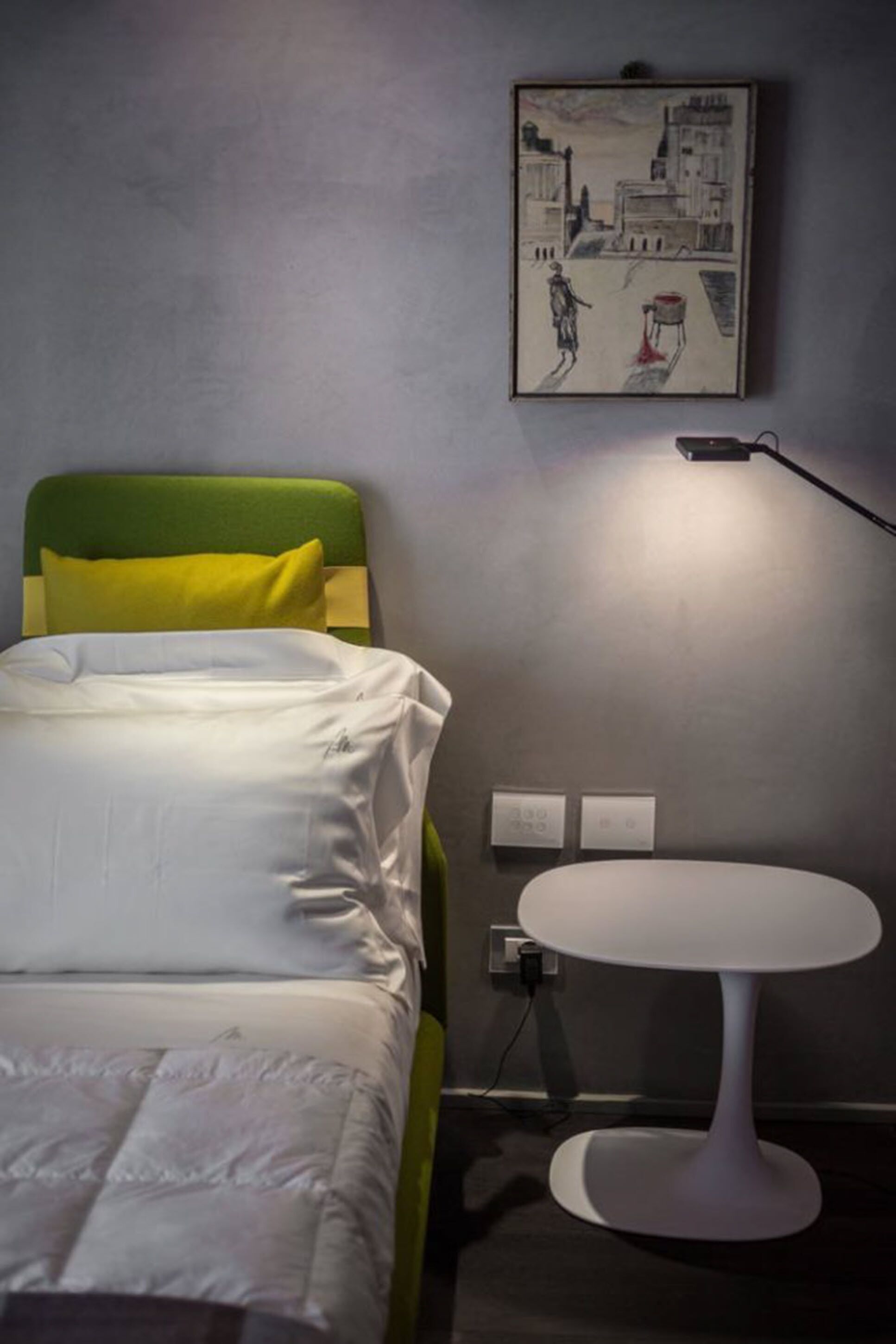 Una habitación de hotel con una cama, un cabecero verde, una mesita de noche blanca, una mesita redonda pequeña y una lámpara fijada a la pared.