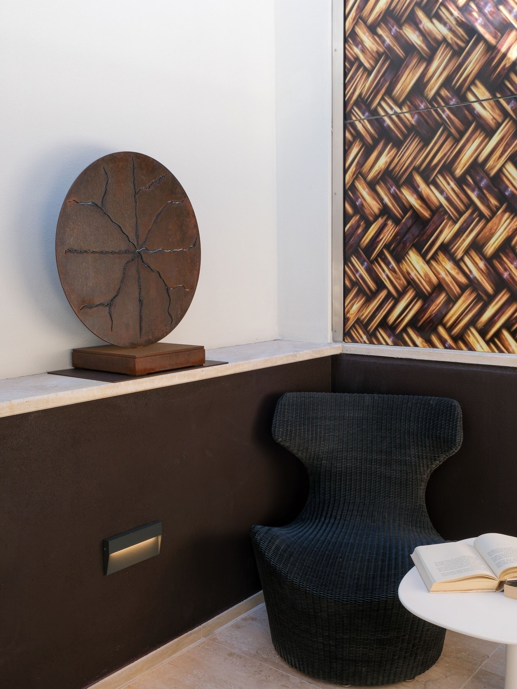 A modern room with a round, cracked wooden wall art piece, a wicker chair, and a small table with an open book.
