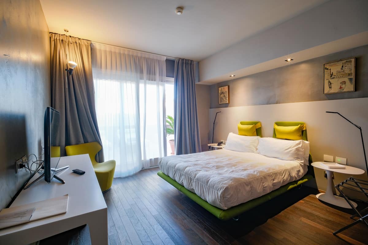 A modern hotel room with a bed, desk, chair, and a view of the outdoors.