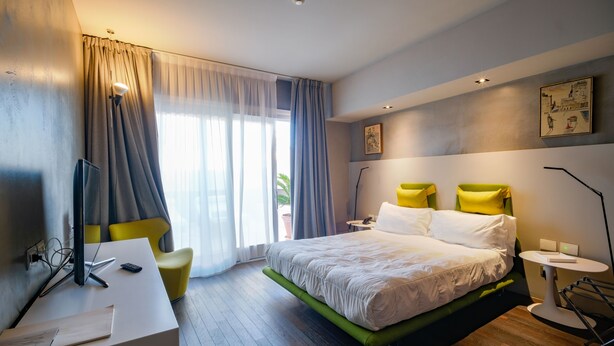 A modern hotel room with a bed, desk, chair, and a view of the outdoors.