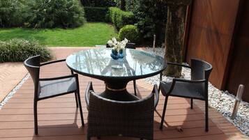 Mesa redonda de vidrio con cuatro sillas de mimbre, rodeada por una terraza de madera y vistas a un jardín.