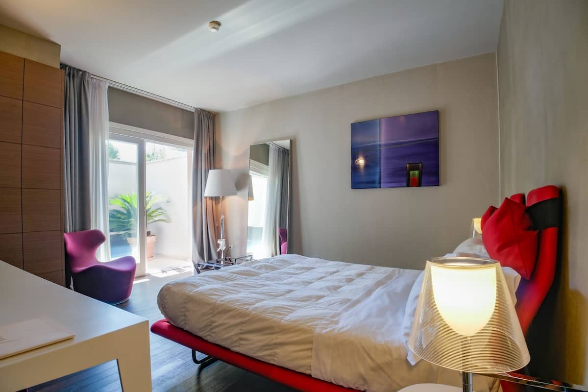 A hotel room with a bed, a desk, a chair, a lamp, a painting, and a window with curtains.