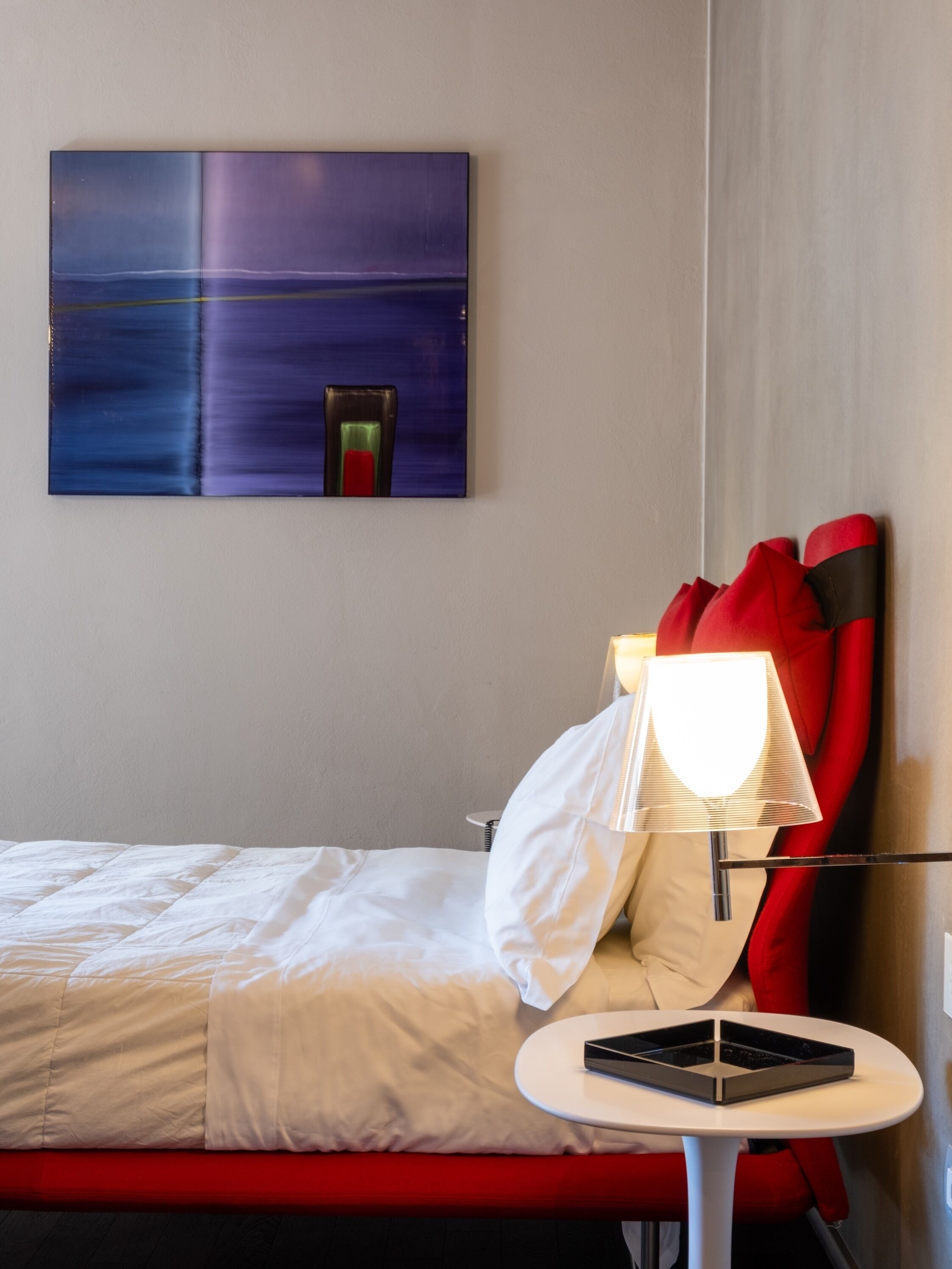 A hotel room with a bed, a table with a lamp, a red chair, and a painting on the wall.