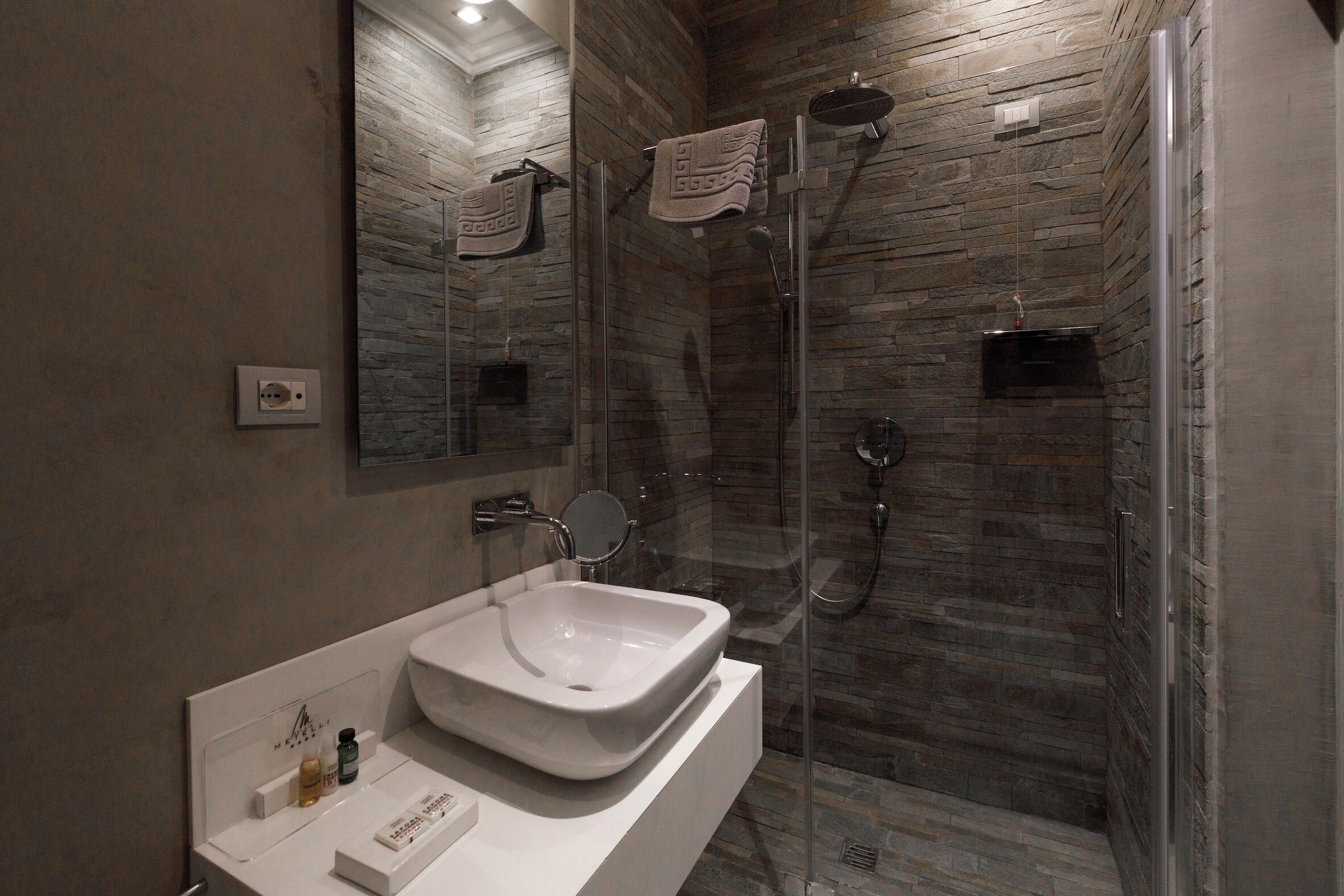 Ein modernes Badezimmer mit Steinwand, einer gläsernen Dusche, einem weißen Waschbecken und einem Spiegel.