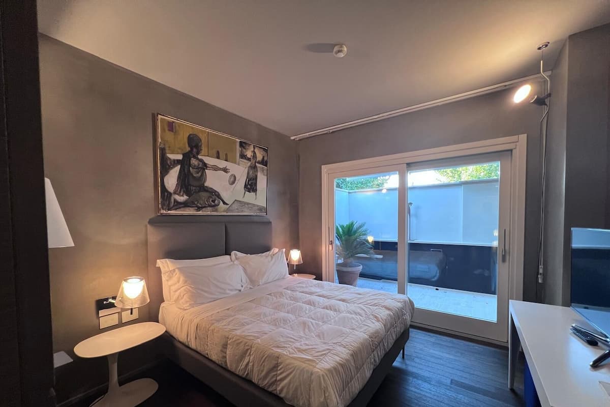 A modern bedroom with a bed, bedside tables, a painting, and a sliding glass door to a balcony.