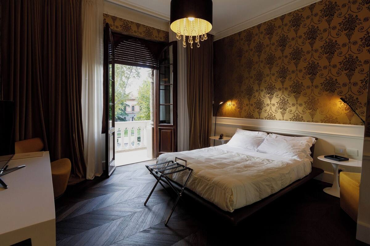 A hotel room with a bed, a desk, a chair, a hanging light fixture, and a view through a window.