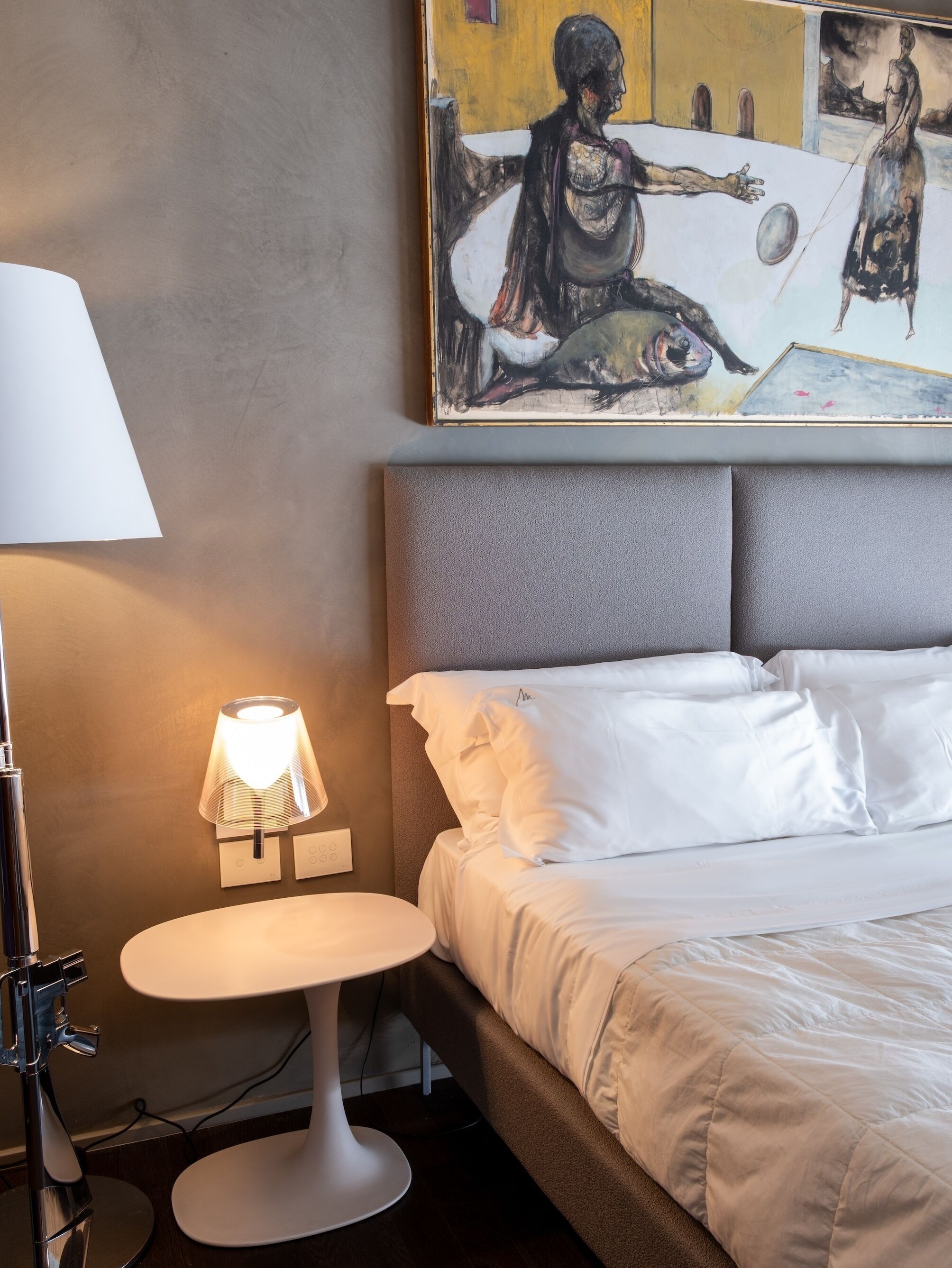 A hotel room with a bed, a bedside lamp, a small table, and a painting on the wall.