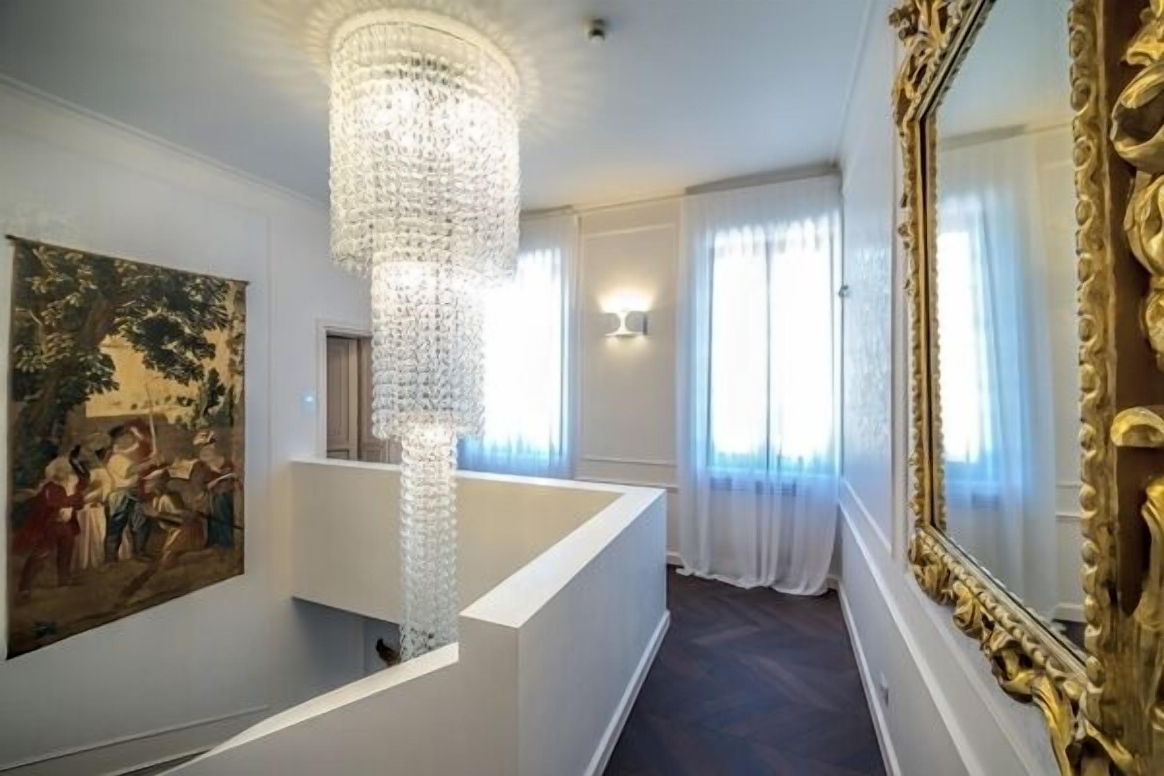 A grand hallway with a tall, ornate chandelier, a large mirror with a golden frame, and a painting on the wall.